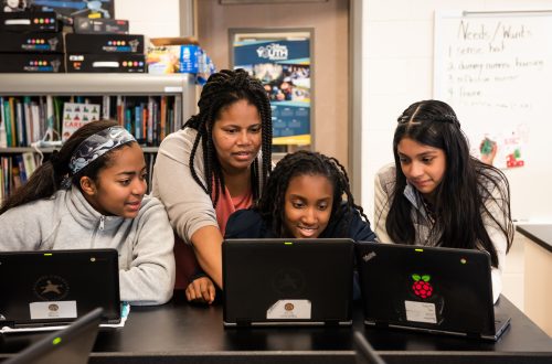 A female computing educator with three female students at laptops in a classroom.
