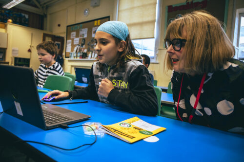 A girl codes at a laptop while a woman looks on during a Code Club session.