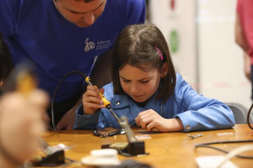 A young girl solders something at a worktop while a man looks over her shoulder.