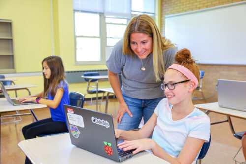 In a classroom, a teacher and a student look at a computer screen while the student types on the keyboard.