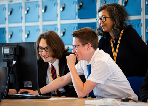 Two teenage boys do coding at a shared computer during a computer science lesson while their woman teacher observes them.