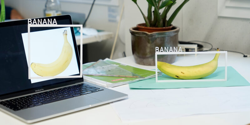 An image demonstrating that AI systems for object recognition do not distinguish between a real banana on a desk and the photo of a banana on a laptop screen.