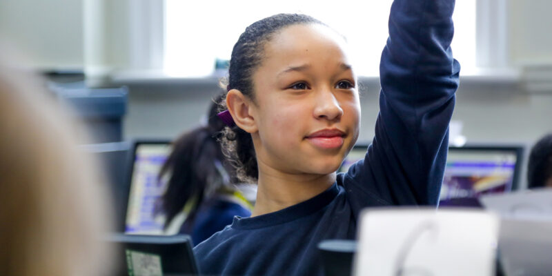 In a computing classroom, a smiling girl raises her hand.
